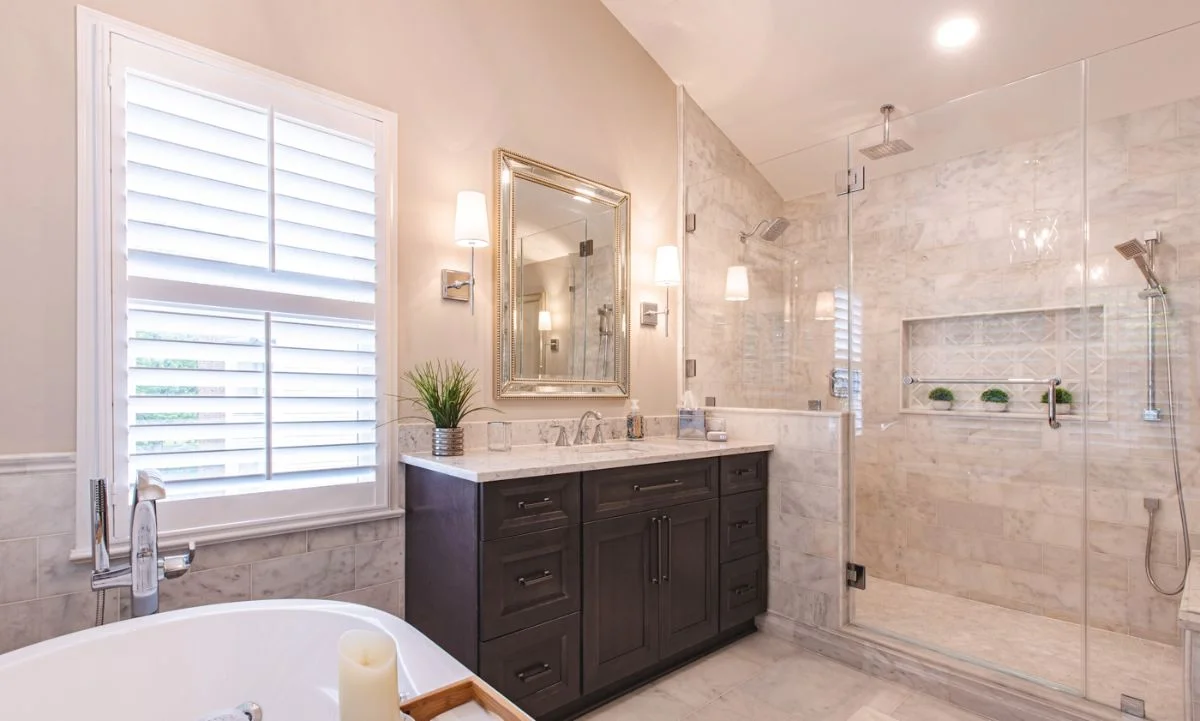Dark wood vanity next to window with plantation shutters and glass shower entrance in Ashburn VA bathroom.