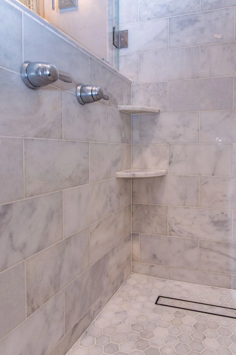 Marble shower corner with soap shelves, chrome valves, and hexagon mosaic floor with linear drain in Ashburn VA.