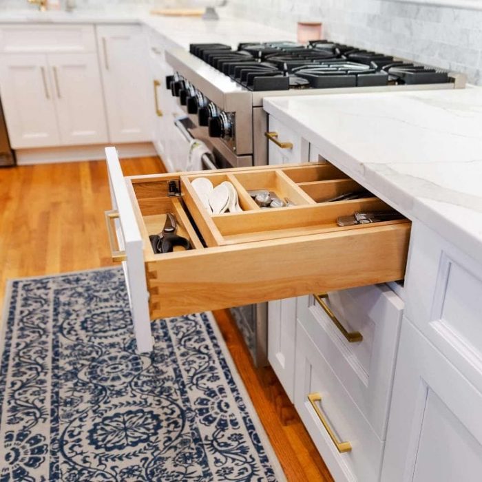 Open white Shaker drawer featuring a wooden tiered cutlery organizer, dovetail construction, and gold hardware next to a gas range.