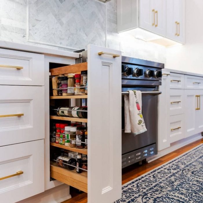 Pull-out wooden spice rack cabinet next to a stainless steel range in a white Shaker kitchen.