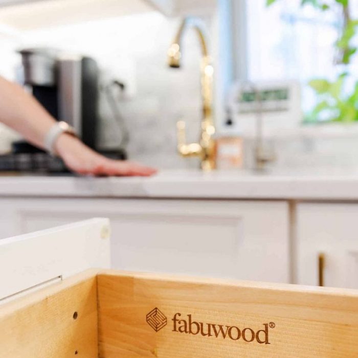 Close-up of Fabuwood logo on a wooden drawer box in a white Shaker kitchen with gold accents.