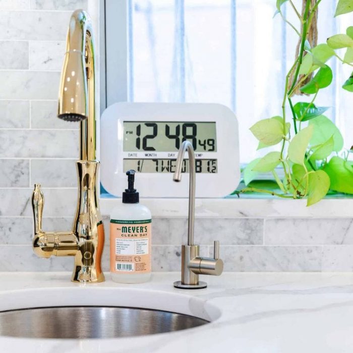 Close-up of a gold bar faucet and round stainless steel prep sink with marble subway tile backsplash and trailing plants.
