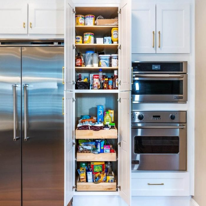Integrated kitchen wall with a stainless steel refrigerator, stacked wall ovens, and a white Shaker pantry with pull-out wooden trays.