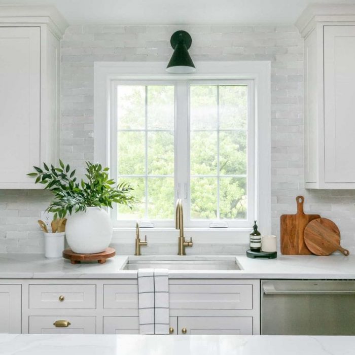 Kitchen sink area featuring a window with black sconce, gold faucet, stainless steel dishwasher, and textured white tile backsplash.