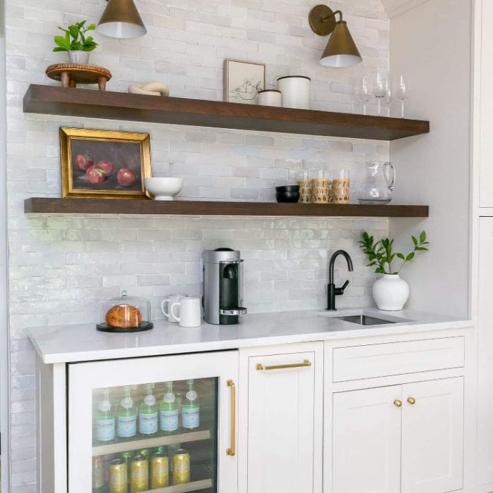 Modern wet bar with floating wood shelves, white textured tile backsplash, beverage fridge, and brass wall sconces.