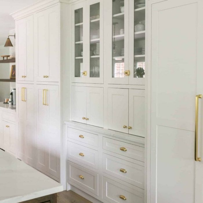 Floor-to-ceiling white Shaker storage wall featuring glass display cabinets, pantry drawers, and mixed gold hardware.