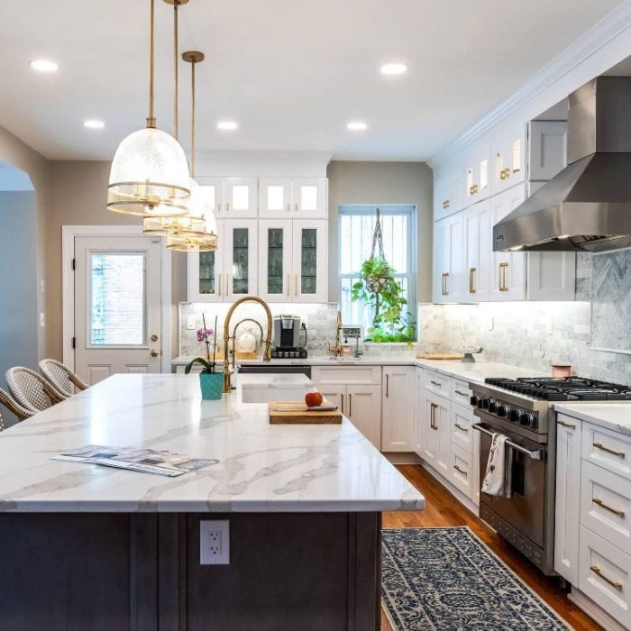 Luxury transitional kitchen featuring a dark grey island, white Shaker cabinets with gold hardware, and marble herringbone backsplash.