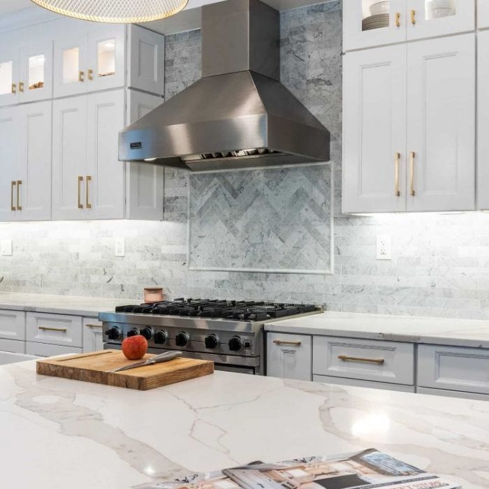 Kitchen island detail with USA Cabinet Store catalog, professional stainless steel vent hood, and herringbone marble backsplash.