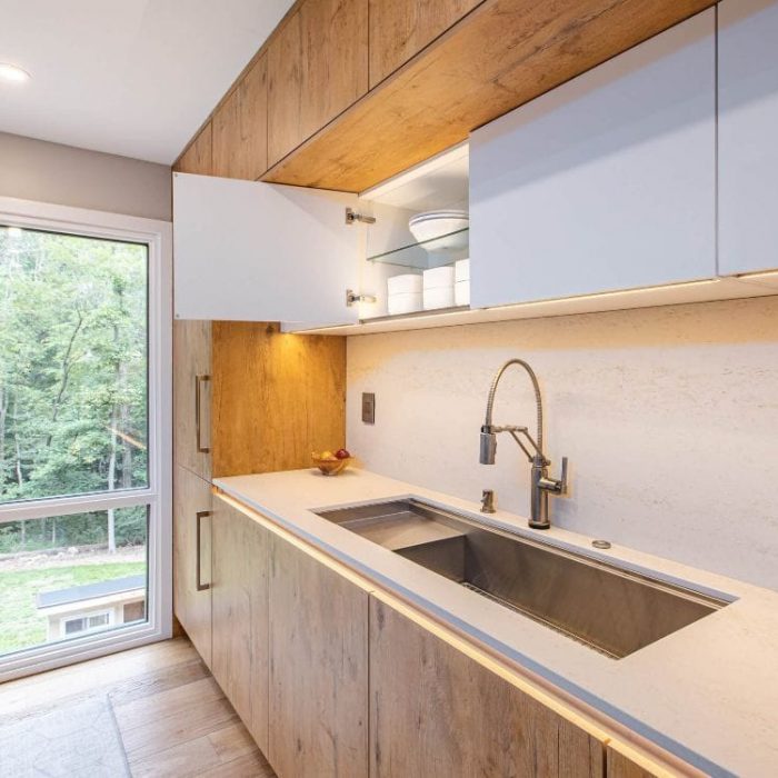 Modern kitchen detail featuring open white glossy cabinets, wood-grain base units, a stainless steel sink, and a forest view window.