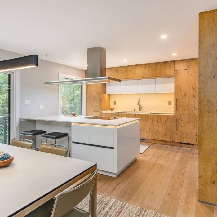 Modern open-concept home with a white glossy island, natural wood cabinetry, linear lighting, and large windows overlooking a forest.