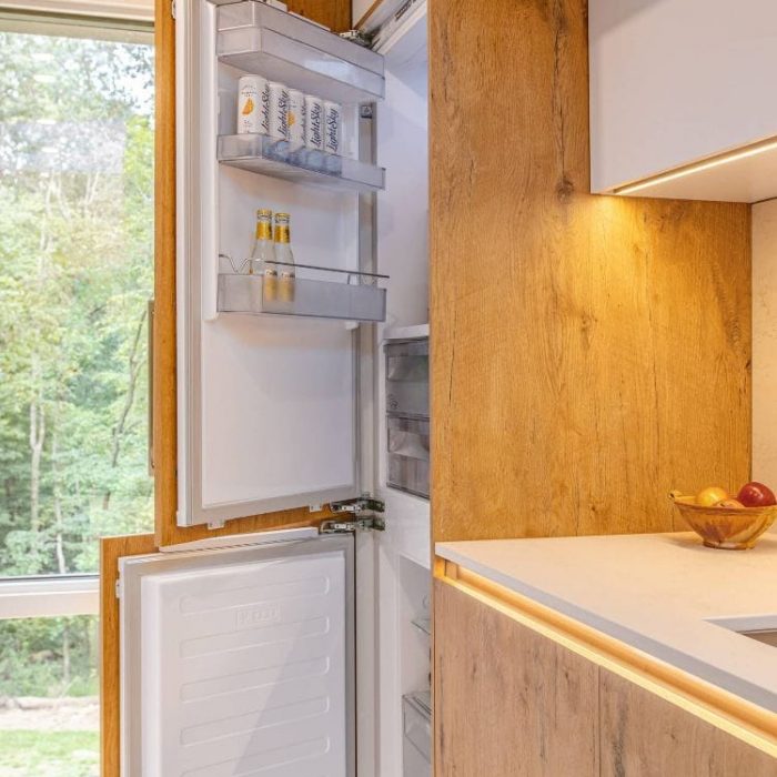 Integrated panel-ready refrigerator concealed behind natural wood cabinetry with LED accent lighting and forest views.